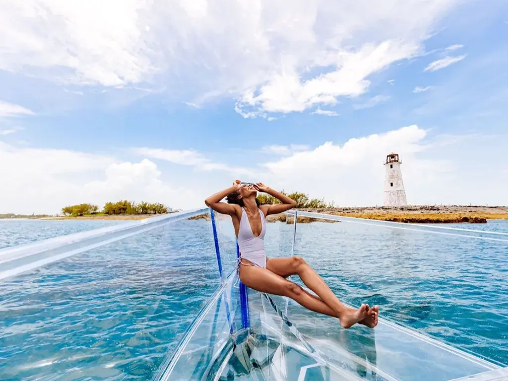 A traveler lounges on the bow of a boat on clear blue water, with a lighthouse on the horizon.