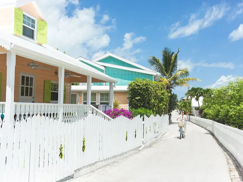 Pastel cottages behind white picket fences line a sunny lane beneath palm trees.