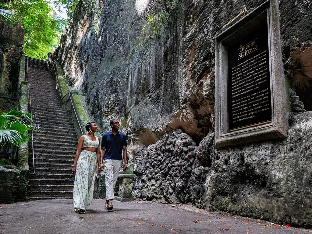 A couple walks through the limestone passage of the Queen's Staircase in Nassau, beside its plaque.