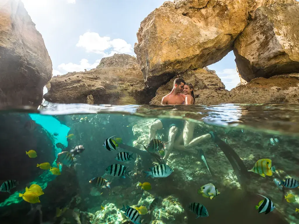 A split view of a couple in a rock arch above the water and reef fish swimming below it in The Exumas.