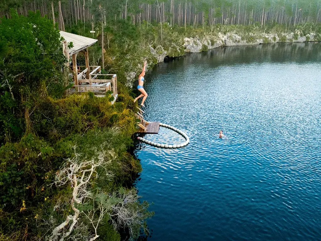 Swimmers leap from a small platform into a forest-ringed blue hole on Andros.