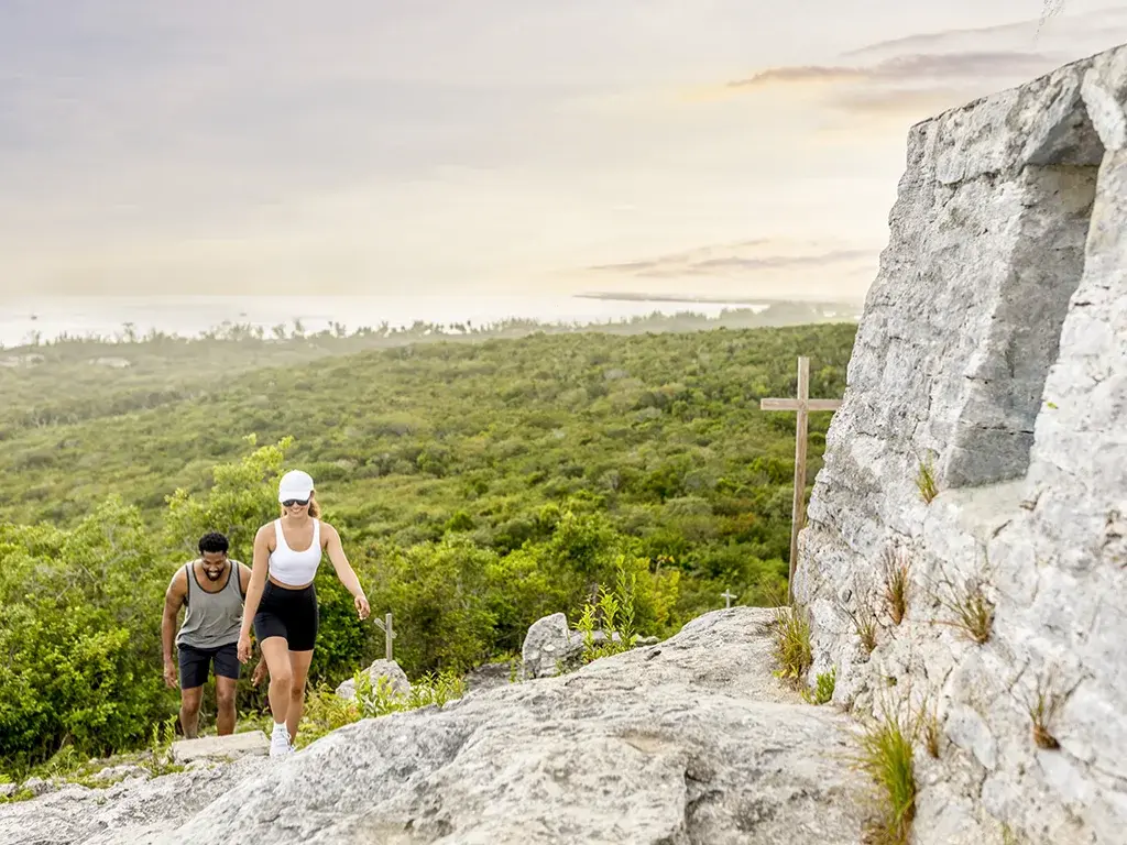 Hikers climb a rocky hilltop path toward a stone cross on Cat Island.