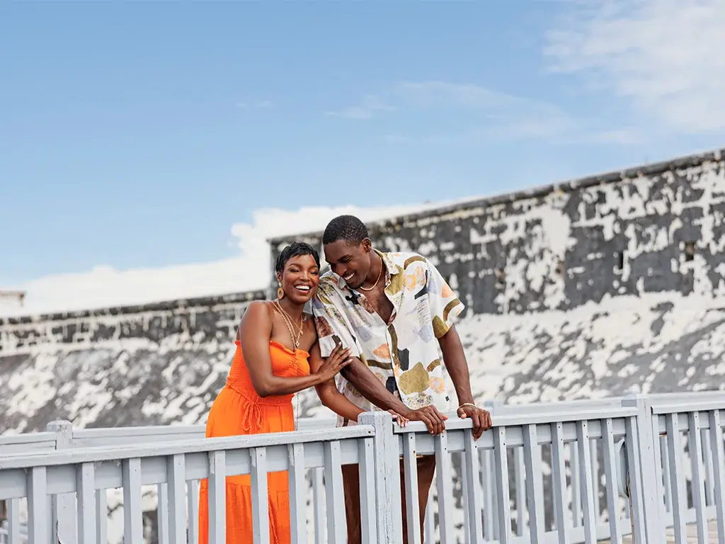 A couple stands at a railing in front of a weathered white fort wall.
