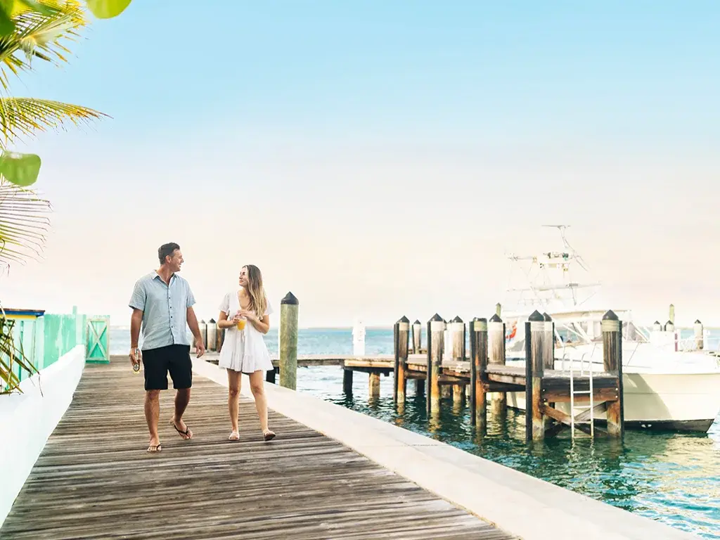 A couple walks along a marina dock past boats.