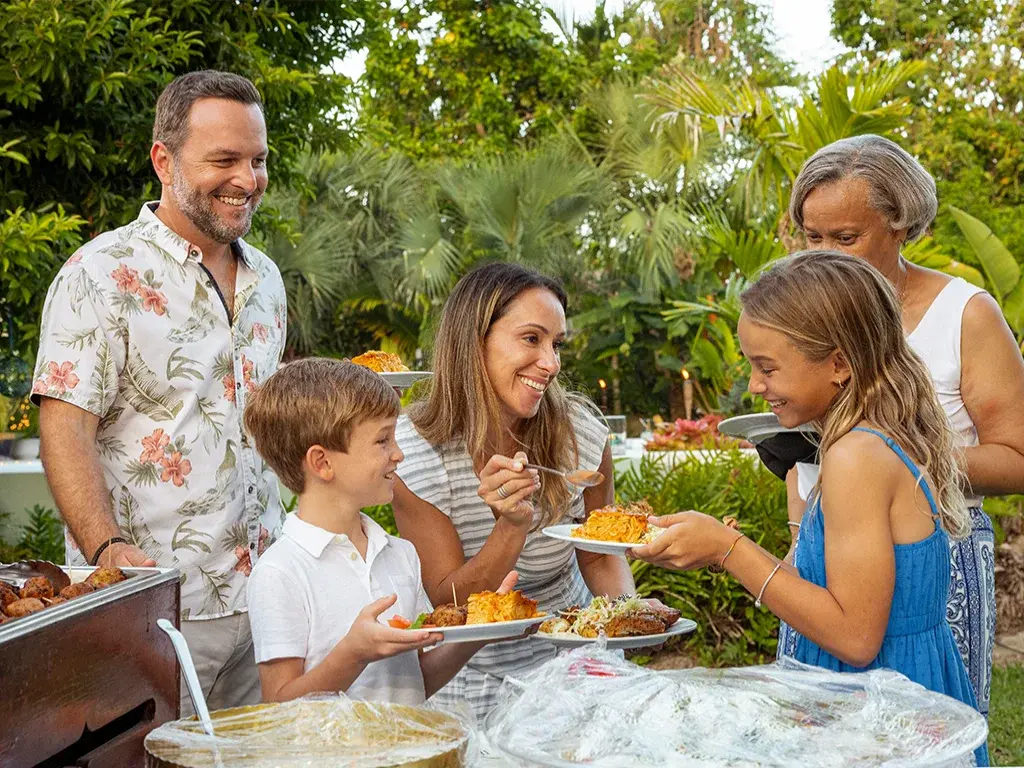A host serves a visiting family at an outdoor buffet.