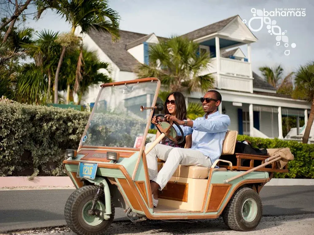A couple drives a golf cart past a white colonial house.