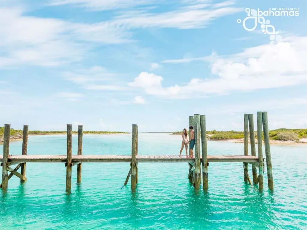 Two travelers stand on a rustic dock over turquoise water.